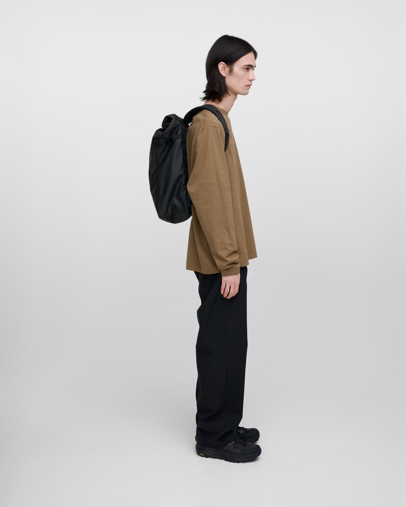 Person wearing a brown jacket and black pants with a black backpack on a white background.
